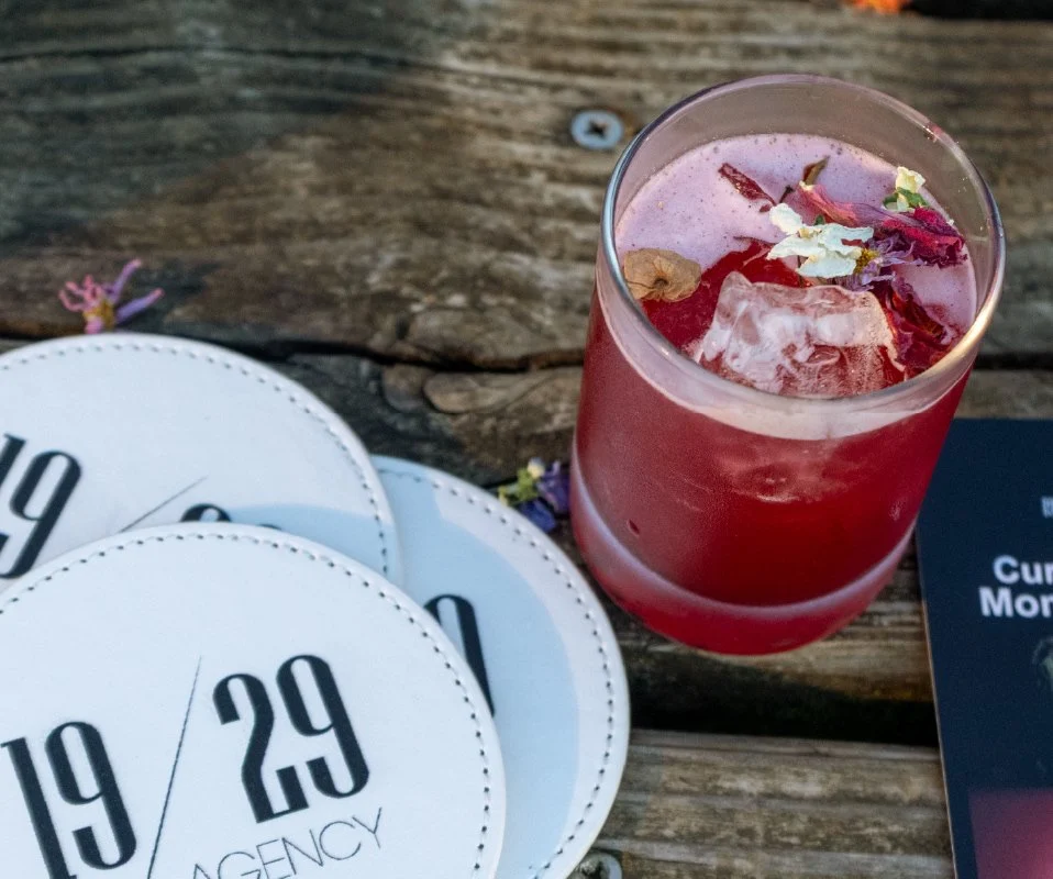 Red cocktail beside 19/29 Agency coasters on a wooden table