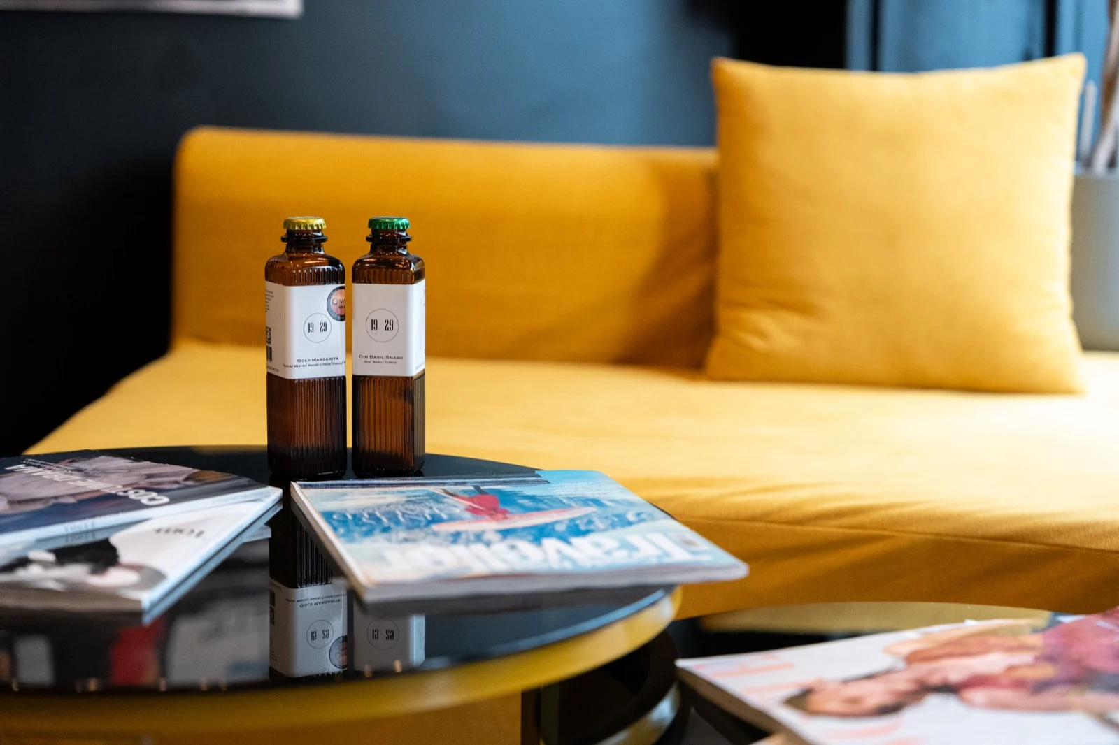 Two 19/29 bottles on a table beside a yellow sofa, with magazines