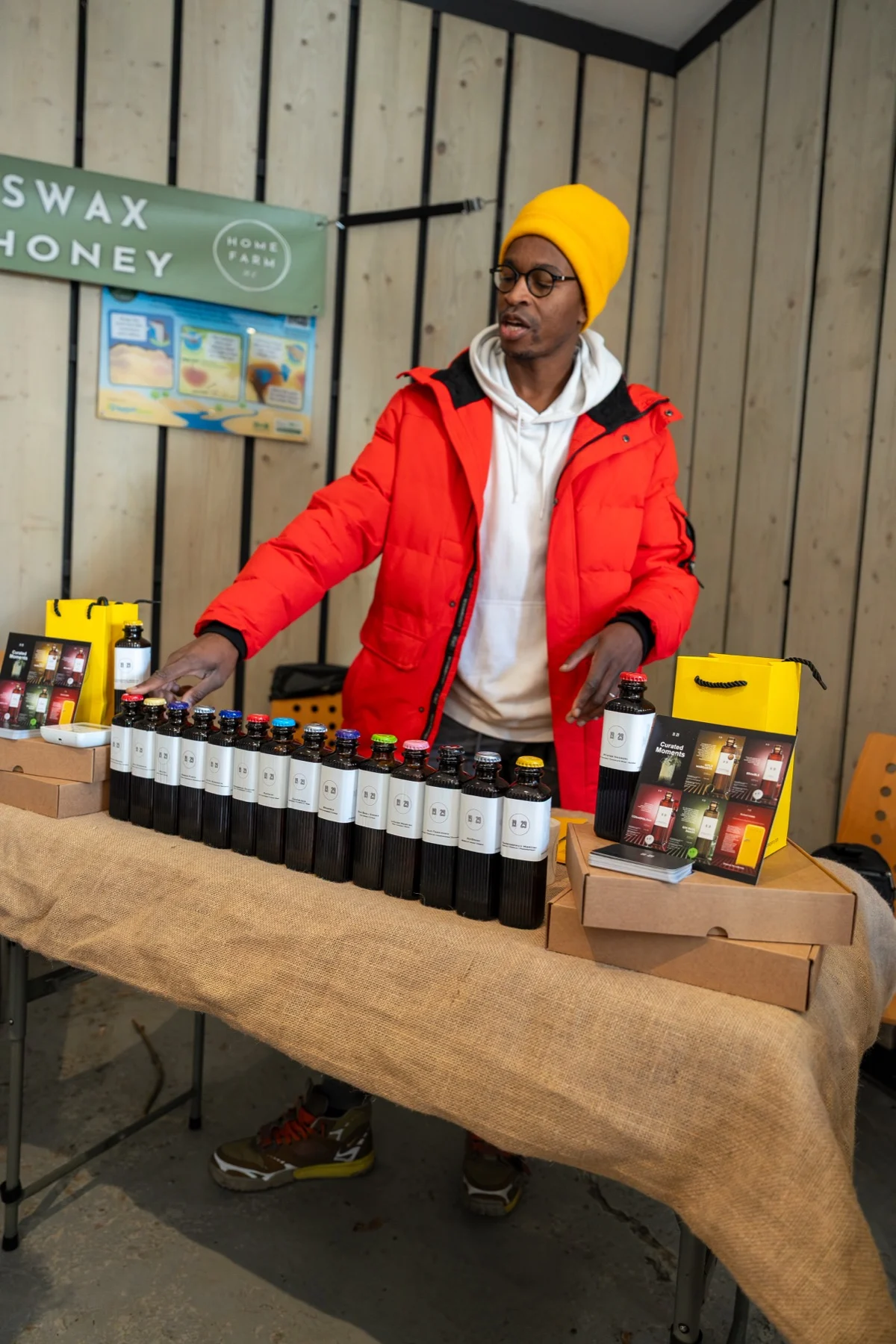 A bartender in a yellow beanie and red jacket behind a row of 19/29 bottles