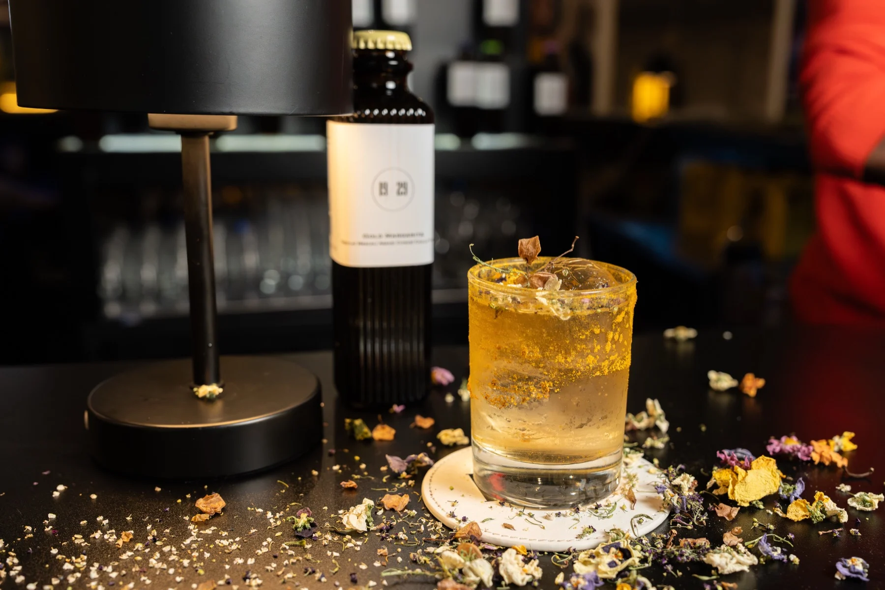 A golden cocktail with dried flowers beside a 19/29 bottle